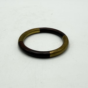 May include: A circular bangle bracelet featuring alternating segments of dark brown wood and brushed gold-tone metal. The smooth, rounded band has a polished finish on the wooden sections and a textured, metallic appearance on the gold-colored parts.