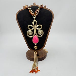 Op de afbeelding: Een goudkleurige statement ketting met een grote, sierlijke hanger met een roze druppelvormige steen. De ketting heeft een kwastje van gouden kettingen en oranje kralen. De ketting wordt tentoongesteld op een houten sieradenstandaard.