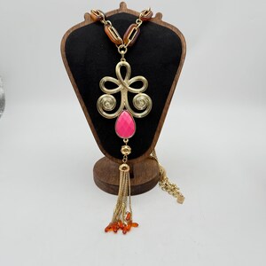 Op de afbeelding: Goudkleurige statement ketting met een grote roze druppelvormige hanger en een kwast van gouden kettingen en oranje kralen. De ketting heeft een decoratief gouden filigraan ontwerp en bruine en gouden schakels.