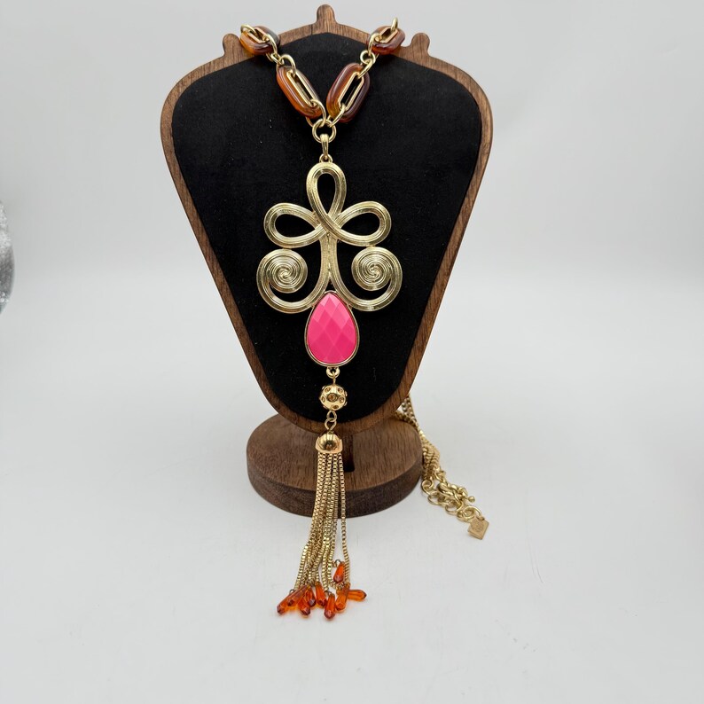 Op de afbeelding: Goudkleurige statement ketting met een grote roze druppelvormige hanger en een kwast van gouden kettingen en oranje kralen. De ketting heeft een decoratief gouden filigraan ontwerp en bruine en gouden schakels.