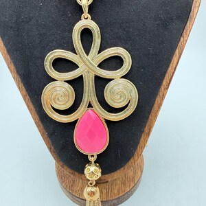 Op de afbeelding: Goudkleurige hangerketting met een groot, sierlijk ontwerp met wervelende patronen en een druppelvormige, gefacetteerde roze steen. De hanger hangt aan een fijne ketting en heeft een kleine kwast.