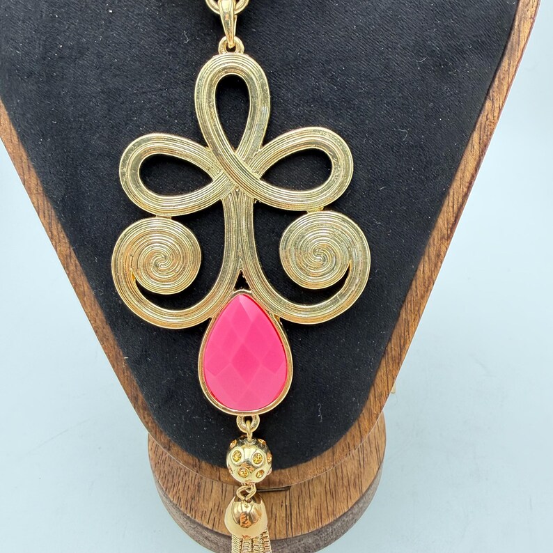 Op de afbeelding: Goudkleurige hangerketting met een groot, sierlijk ontwerp met wervelende patronen en een druppelvormige, gefacetteerde roze steen. De hanger hangt aan een fijne ketting en heeft een kleine kwast.