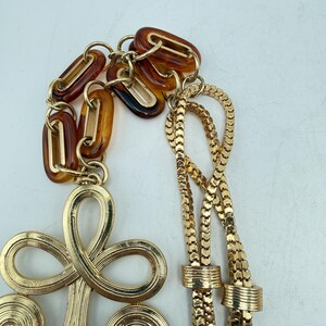 Op de afbeelding: Een goudkleurige ketting met een grote, geluste hanger en een ketting van kleinere, in elkaar grijpende schakels. De ketting heeft een reeks amberkleurige, ovaalvormige accenten. De ketting is gemaakt van een delicaat, geweven ontwerp.