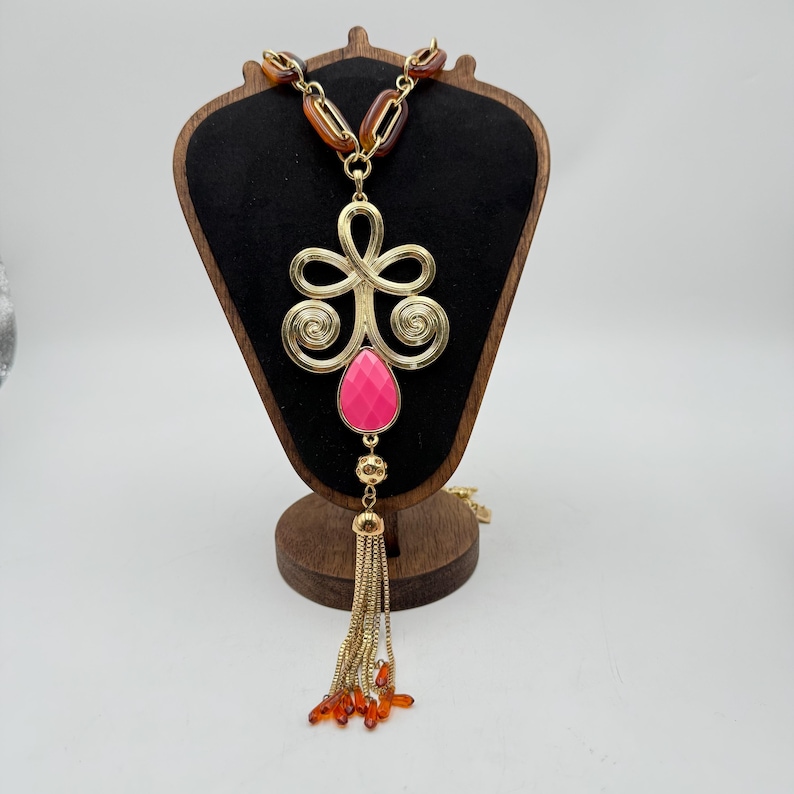 Op de afbeelding: Een goudkleurige statement ketting met een grote, sierlijke hanger met een roze druppelvormige steen. De ketting heeft een kwastje van gouden kettingen en oranje kralen. De ketting wordt tentoongesteld op een houten sieradenstandaard.
