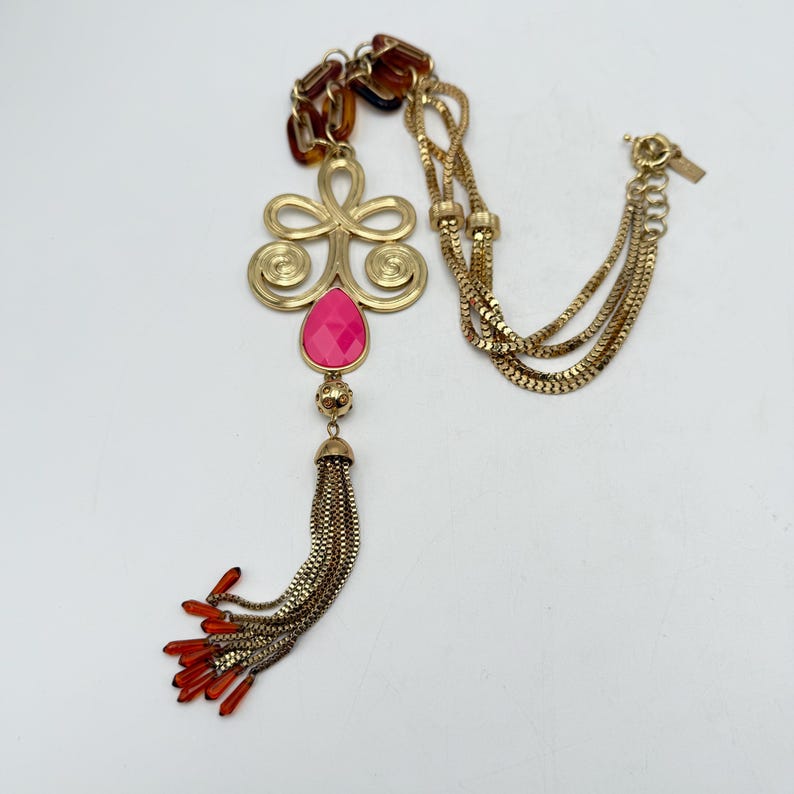 Op de afbeelding: Goudkleurige ketting met een grote hanger met een roze druppelvormige steen en een kwast. De ketting heeft een ketting met een mix van gouden en schildpadkleurige schakels. De kwast heeft oranjerode kralen aan het uiteinde.