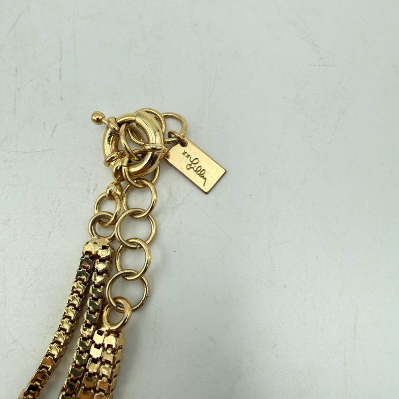 Op de afbeelding: Close-up van een goudkleurige ketting met een toggle sluiting. De ketting heeft meerdere strengen ketting en een klein rechthoekig label met de merknaam. De kettingen hebben een getextureerde, geweven uitstraling.