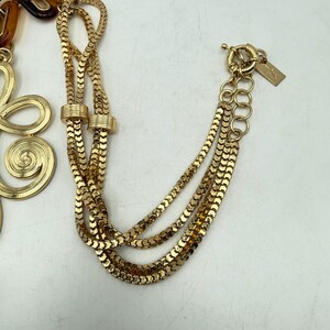 Op de afbeelding: Goudkleurige ketting met meerdere strengen en een decoratief bloemelement. De ketting heeft een karabijnsluiting en een klein label. Het ontwerp omvat een mix van kettingstijlen, waardoor een gelaagd effect ontstaat.