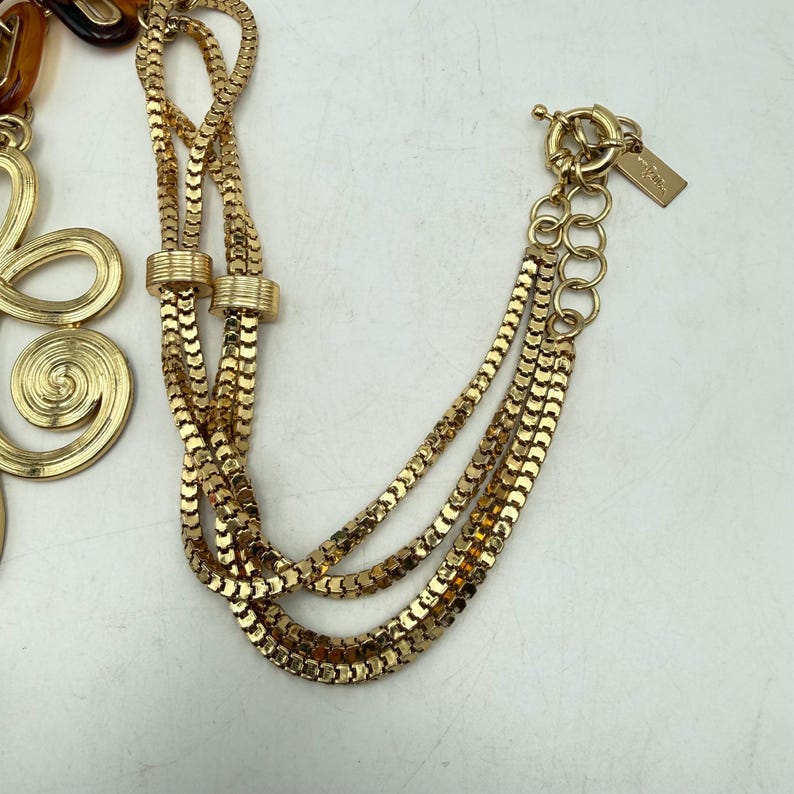 Op de afbeelding: Goudkleurige ketting met meerdere strengen en een decoratief bloemelement. De ketting heeft een karabijnsluiting en een klein label. Het ontwerp omvat een mix van kettingstijlen, waardoor een gelaagd effect ontstaat.