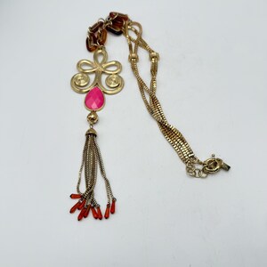 Op de afbeelding: Goudkleurige ketting met een hanger met een roze druppelvormige steen en een kwast van kleine kettingen. De ketting heeft een decoratief bloemmotief en rode accenten. De ketting is een ontwerp met meerdere strengen.