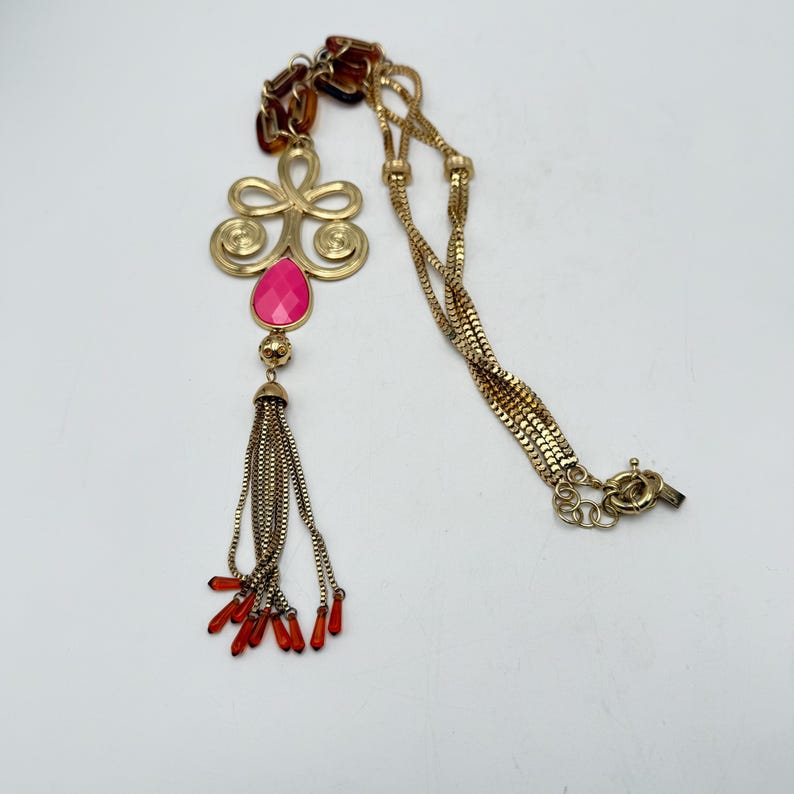 Op de afbeelding: Goudkleurige ketting met een hanger met een roze druppelvormige steen en een kwast van kleine kettingen. De ketting heeft een decoratief bloemmotief en rode accenten. De ketting is een ontwerp met meerdere strengen.