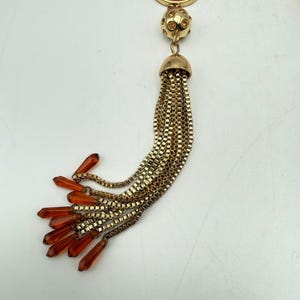 Op de afbeelding: Een goudkleurige hanger met een kwastontwerp. De hanger heeft een ronde bovenkant met een kleine, met studs bezette bol en een goudkleurige, klokvormige dop waaruit meerdere gouden kettingen naar beneden vallen. De kettingen eindigen met oranjerode, druppelvormige kralen.