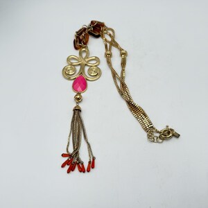Op de afbeelding: Goudkleurige ketting met een hanger met een roze druppelvormige steen en een kwast. De ketting heeft een ketting met een sluiting en een bruine ketting aan de bovenkant.