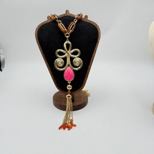 Op de afbeelding: Goudkleurige statement ketting met een grote roze druppelvormige hanger en een gouden kwast. De ketting heeft een decoratief gouden filigraan ontwerp en bruine en oranje accenten. De ketting wordt getoond op een houten sieradenstandaard.