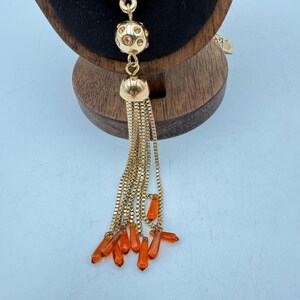 Op de afbeelding: Goudkleurige ketting met een kwastontwerp. De ketting heeft een ronde, met juwelen bezette kraal, een klokvormig element en een kettingkwast met oranje druppelvormige kralen. De ketting wordt gepresenteerd op een houten standaard.