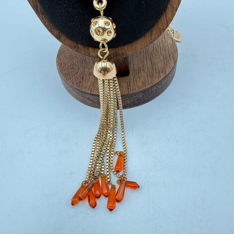 Op de afbeelding: Goudkleurige ketting met een kwastontwerp. De ketting heeft een ronde, met juwelen bezette kraal, een klokvormig element en een kettingkwast met oranje druppelvormige kralen. De ketting wordt gepresenteerd op een houten standaard.