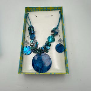 May include: A blue and teal necklace and earring set with a large, round pendant. The jewelry is displayed in a yellow and blue patterned box. The necklace and earrings feature iridescent shell-like elements and beads.