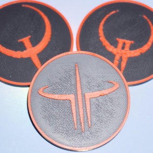 May include: Three circular coasters with a gaming theme. The coasters feature a black or gray background with an orange logo. The logos are stylized and geometric, with a distinct, bold design. The coasters are likely made of a durable material.
