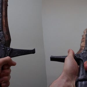 K&ouml;nnte beinhalten: Nahaufnahme einer Person, die ein schwarzes und braunes Schwert mit einem detaillierten Griff und einem Holzgriff h&auml;lt.