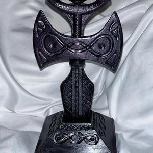 May include: A black metal sculpture of a double-headed axe with intricate details and a square base.