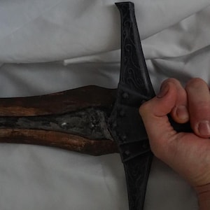 K&ouml;nnte beinhalten: Ein schwarzes Schwert mit einem Holzgriff und einer silbernen Klinge. Das Schwert wird von einer Hand gehalten.