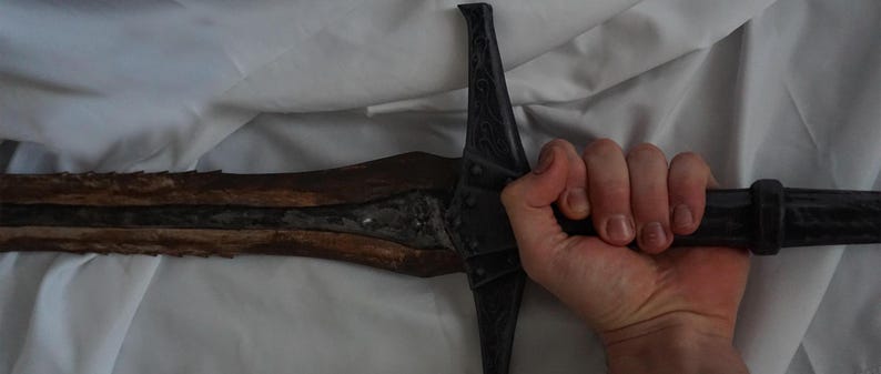 K&ouml;nnte beinhalten: Ein schwarzes Schwert mit einem Holzgriff und einer silbernen Klinge. Das Schwert wird von einer Hand gehalten.
