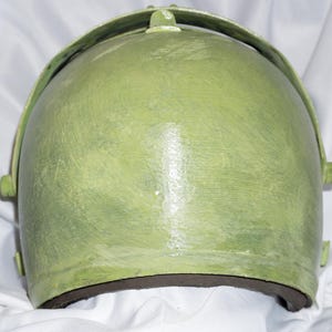 May include: A green helmet with a textured surface and a black rim. The helmet has a curved shape and a raised design on the top.