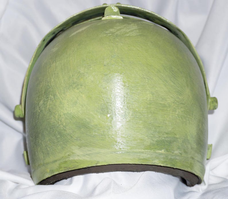 May include: A green helmet with a textured surface and a black rim. The helmet has a curved shape and a raised design on the top.