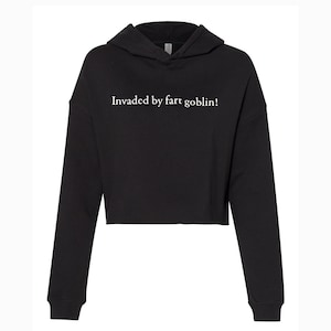 May include: Black cropped hoodie with long sleeves and a hood. The text "Invaded by fart goblin!" is printed in white across the chest. The hoodie is made of a soft material.