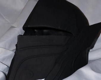 Könnte beinhalten: Ein schwarzer Helm mit spitzer Oberseite und dekorativen Seitenelementen. Der Helm hat ein Visier und liegt auf einer weißen Oberfläche. Die Oberfläche des Helms hat geprägte Muster.