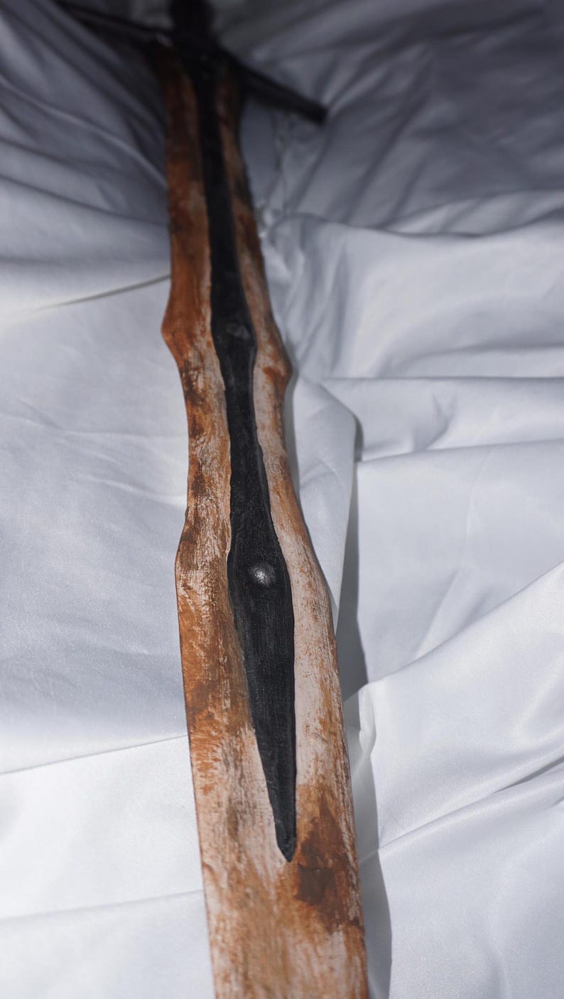 K&ouml;nnte beinhalten: Ein Holzschwert mit einer schwarz lackierten Klinge. Das Holz ist in einem braunen und wei&szlig;en Farbschema lackiert, was ihm ein verwittertes Aussehen verleiht.