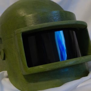 May include: A green military-style helmet with a dark visor. The helmet has a textured surface and a rounded shape.