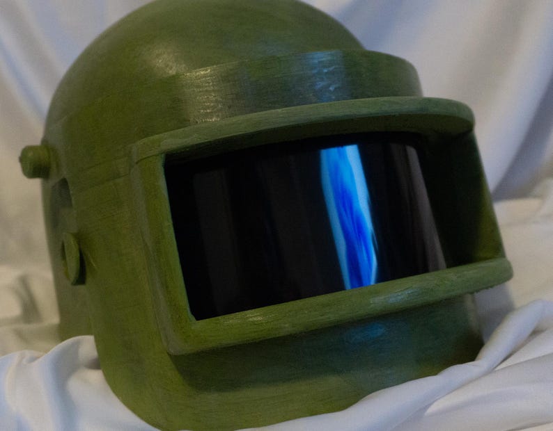 May include: A green military-style helmet with a dark visor. The helmet has a textured surface and a rounded shape.