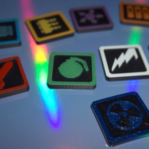 May include: A set of nine square game tiles with different colored borders and black and white symbols. The symbols include a rocket, a bomb, a lightning bolt, a skull, a building, a chain link, a hand, a crown, and a radiation symbol.