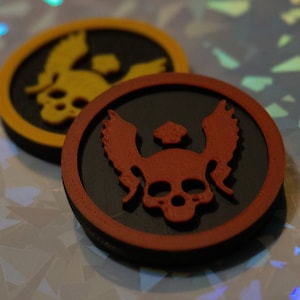 Puede incluir: Dos fichas redondas impresas en 3D, negras y rojas, con un diseño de calavera y alas. La ficha superior tiene una calavera y alas rojas, mientras que la ficha inferior tiene una calavera y alas amarillas.