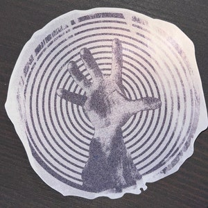 May include: A sticker featuring a grayscale illustration of a hand with fingers extended upwards, set against a backdrop of concentric circular lines. The sticker has a white border and a slightly irregular shape.
