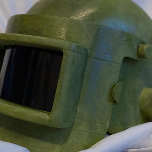 May include: A green military-style helmet with a visor and a textured surface.