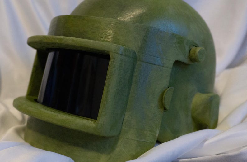 May include: A green military-style helmet with a visor and a textured surface.