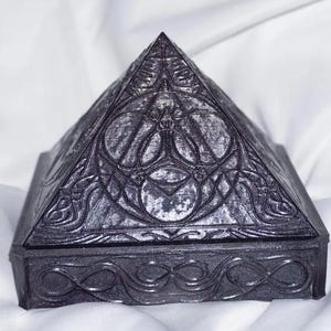 Może przedstawiać: Czarne pudełko w kształcie piramidy z misternymi rzeźbami i srebrzystym wykończeniem. Pudełko ma ozdobną ramkę wokół górnej i dolnej części.