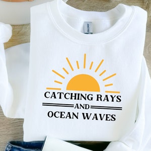 May include: White sweatshirt with the text "CATCHING RAYS AND OCEAN WAVES" in black, with a yellow and orange sun graphic. The sweatshirt is a crew neck style.