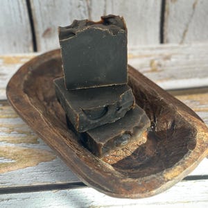 May include: Three dark brown soap bars stacked in a rustic wooden bowl. The soaps have a textured surface and a natural, handmade appearance. The bowl is oval-shaped with a worn, weathered finish.