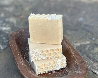 May include: Three beige soap bars stacked in a wooden bowl. The top of each bar has a honeycomb pattern and a decorative edge. The bowl is a dark brown color and sits on a gray stone surface.
