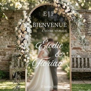 Peut inclure: Une arche de mariage en métal doré décorée de roses blanches et de feuillage. Le texte indique E | F, BIENVENUE A NOTRE MARIAGE, Élodie & Florian, et 17 Juillet 2027. L'arche est placée en extérieur devant un mur en pierre et un jardin.