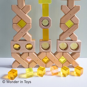 以下が含まれることがあります： カラフルな半透明の幾何学模様の入った木製ブロックのセット。ブロックは3つの塔に配置されており、中央の塔が最も高い。ブロックは天然木で作られており、滑らかな仕上がりです。半透明の形状は、黄色、オレンジ、ピンクの色合いです。