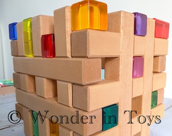 May include: A wooden toy building set with colorful translucent blocks. The blocks are stacked in a tower shape and are made of natural wood. The set includes a variety of colors, including red, yellow, blue, green, and purple. The blocks are designed to be used for creative play and learning.