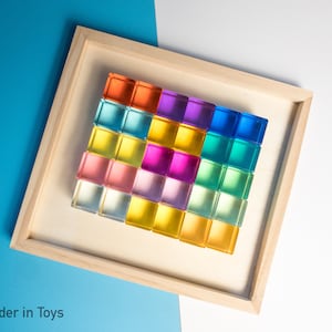 May include: A wooden frame holds a colorful display of 25 clear glass blocks. The blocks are arranged in a grid pattern and are stacked in a rainbow order, starting with red and ending with green.