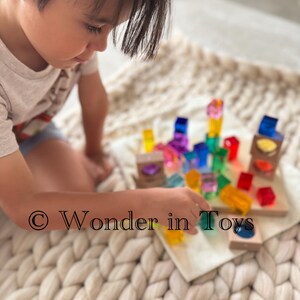 Könnte beinhalten: Ein Kind spielt mit bunten, transparenten Bausteinen und Holzstrukturen auf einem Holztablett. Die Blöcke haben verschiedene Formen und Farben. Der Text "Wonder in Toys" ist sichtbar.