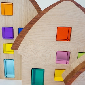 Könnte beinhalten: Holzspeilzeughäuser mit bunten, durchsichtigen Fenstern. Die Häuser sind in einer Reihe angeordnet, wobei das größte Haus hinten und das kleinste Haus vorne steht. Die Fenster sind in verschiedenen Farben gehalten, darunter Orange, Lila, Gelb, Blau, Grün, Rosa und Rot.
