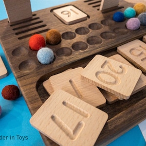 May include: Wooden toy with a number board and colorful felt balls. The board has a grid of holes and a separate area for number tiles. The number tiles are arranged in a stack, with the numbers 1, 2, and 0 visible. The felt balls are in various colors, including red, blue, and yellow.
