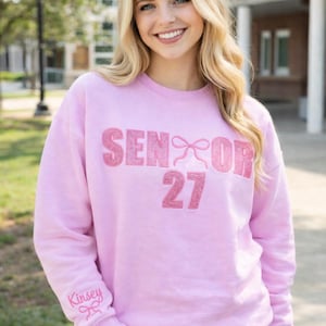 May include: A light pink sweatshirt with the text "SENIOR 27" in a glittery pink font. The word "SENIOR" has a bow design. The sleeve has the name "Kinsey" in a cursive font. The person is wearing blue jeans.