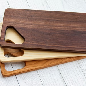May include: Three wooden cutting boards stacked on top of each other. The top board is dark brown, the middle board is light brown, and the bottom board is a reddish brown. All three boards have a unique cutout handle.